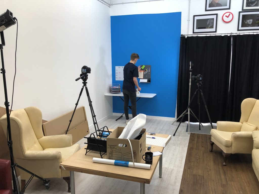 A person stands at a wall-mounted desk with coloured folders in a studio, surrounded by tripods, cameras, cream armchairs, a box on a coffee table, and black curtains, with framed photos and clocks on the wall.