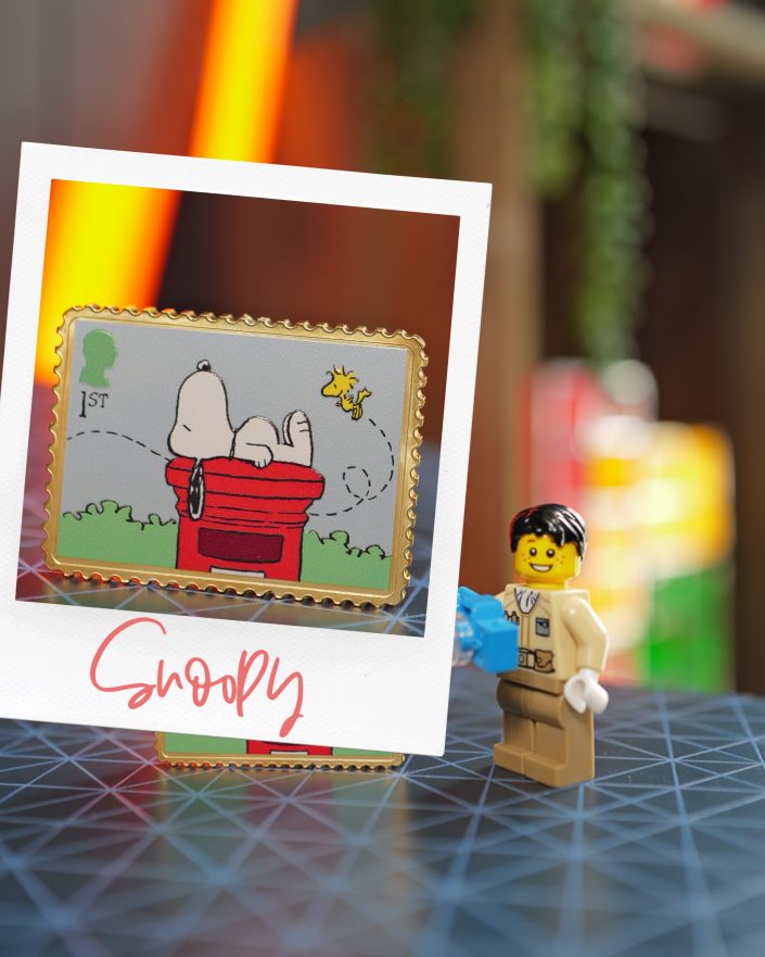 A photo shows a Snoopy pin featuring Snoopy on his red kennel with Woodstock, framed by a white border labelled Snoopy. A smiling LEGO minifigure stands beside it on a patterned surface.