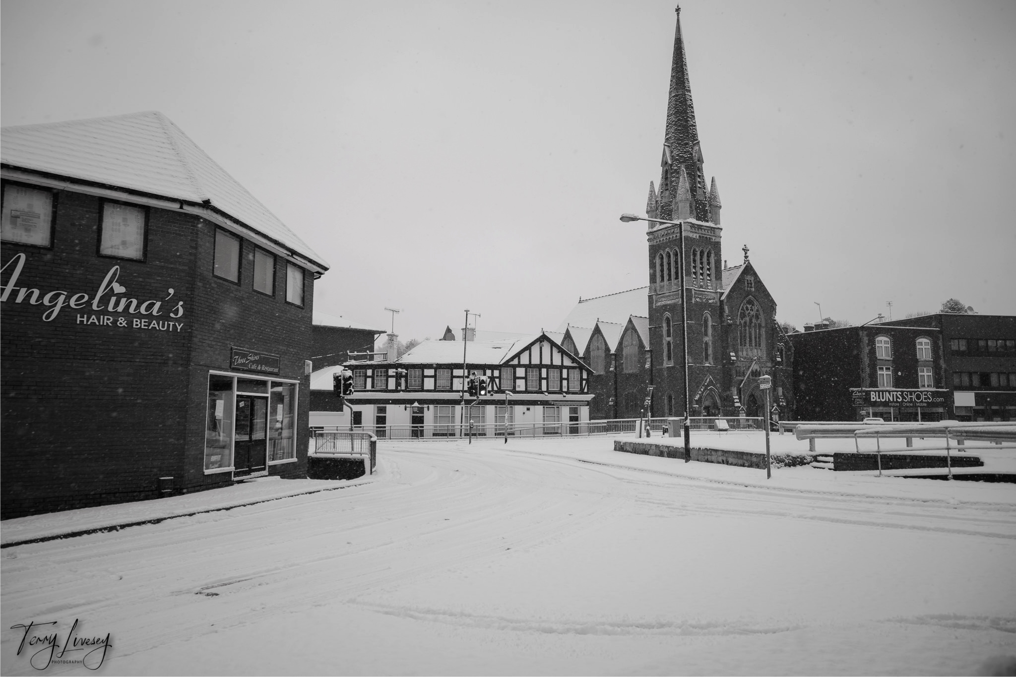 A snowy street scene in Kidderminster shows a church with a tall steeple, shopfronts including Angelinas Hair & Beauty, and visible tracks in the snow. The quiet atmosphere is beautifully captured in this photograph with no people or traffic present.