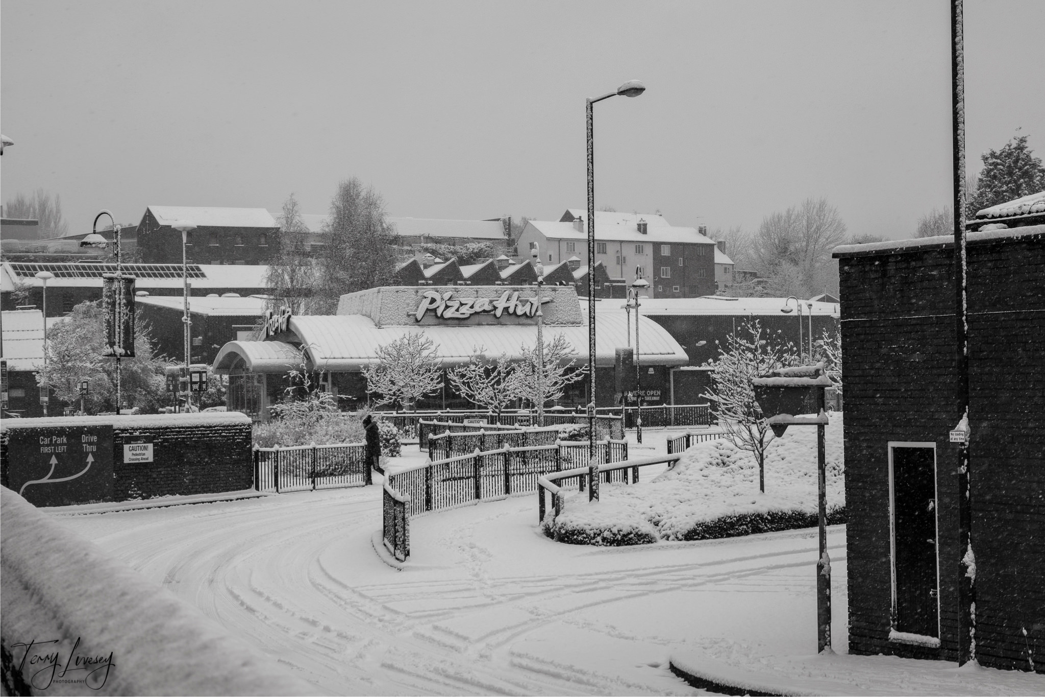 A snowy street in Kidderminster curves past a Pizza Hut restaurant surrounded by trees and buildings. Snow blankets the ground, rooftops, and trees as a few people walk on the pavements under overcast skies, perfect for winter photography.