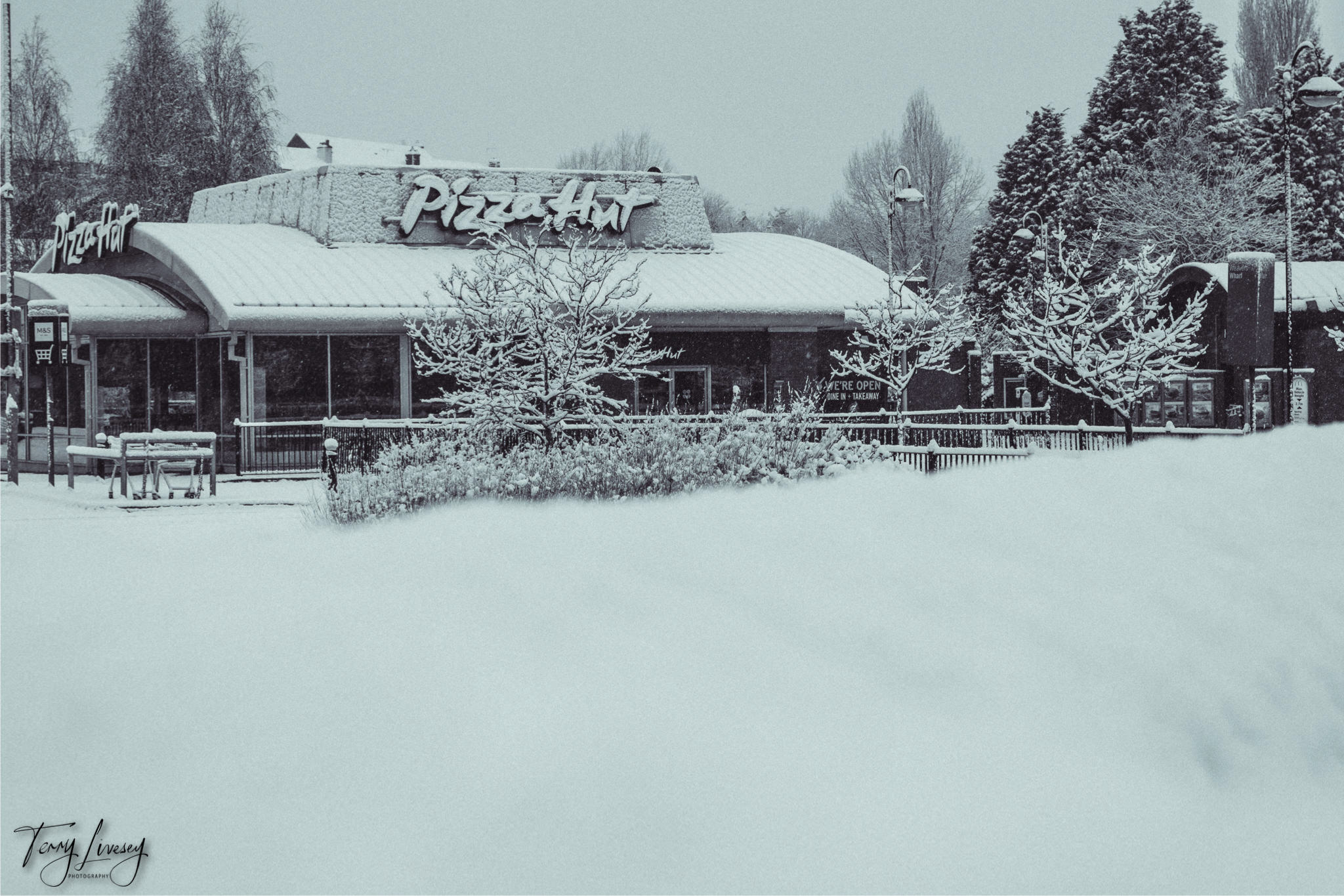 A Pizza Hut restaurant in Kidderminster is covered in snow, with trees, bushes, and the ground blanketed in white. The quiet winter photography scene features bare trees and no visible people.
