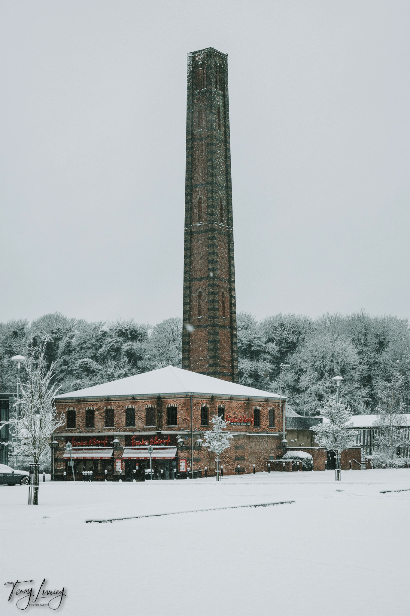 A tall brick chimney rises above a historic Kidderminster building with snowy roofs and surroundings. Snow-covered trees stand behind the structure, beautifully captured in this winter photography under an overcast sky.