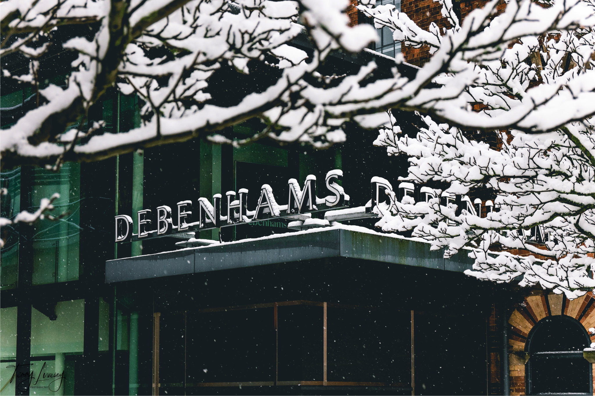 Snow-covered tree branches frame the entrance of a Debenhams store in Kidderminster, with the store’s sign partially visible. Snow is gently falling, adding a wintry atmosphere to this striking piece of urban photography.
