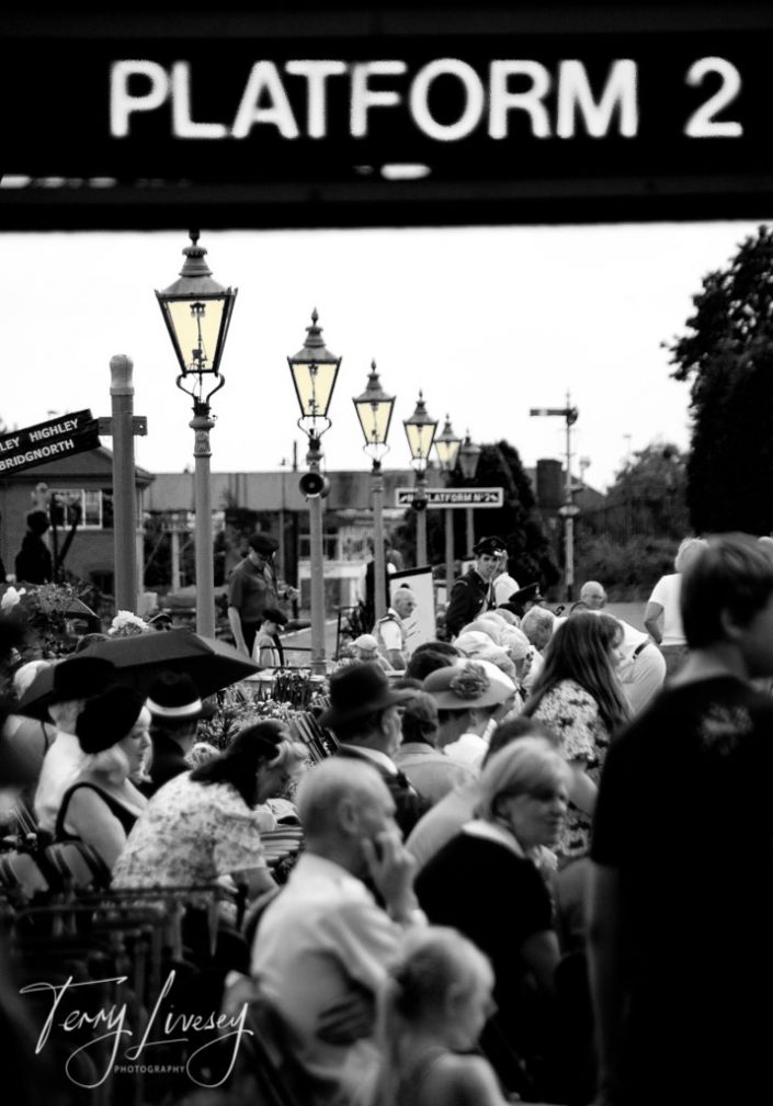 Black-and-white photo of a crowded train platform labelled Platform 2, with many people sitting and standing beneath vintage street lamps. The atmosphere appears busy and nostalgic.