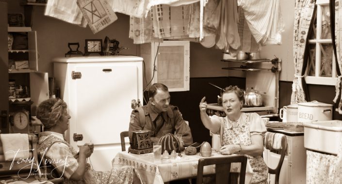 Three people in vintage clothing sit around a kitchen table, talking and laughing. The 1940s-style room has patterned tablecloths, a large fridge, shelves with kitchenware, and a cactus on the table.