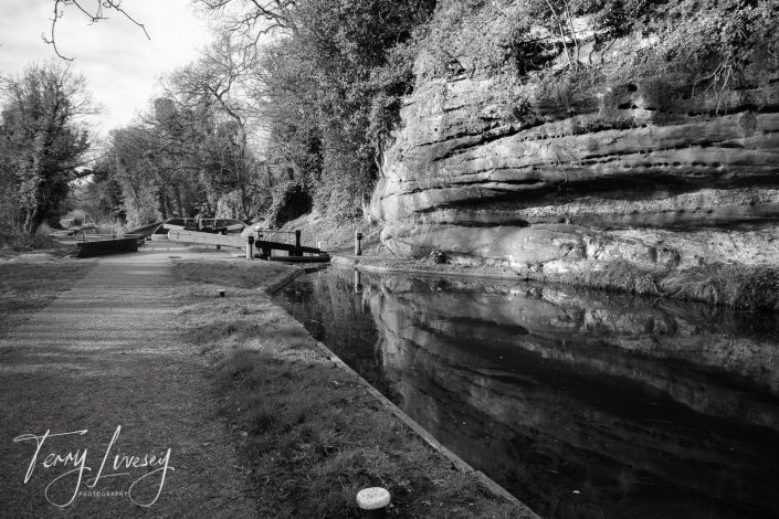Black and white photo of a canal with a steep rock face on the right, calm water reflecting the rock, towpath on the left, trees in the background, and a signature Tony Ivesey Photography in the bottom left corner.