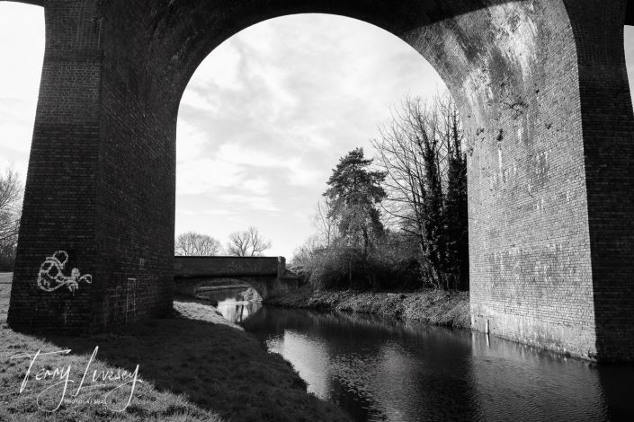 Black-and-white photo of a canal running beneath a large brick arch bridge, with another smaller bridge in the background. Trees and grass line the water’s edge. The image features a signature in the lower left corner.
