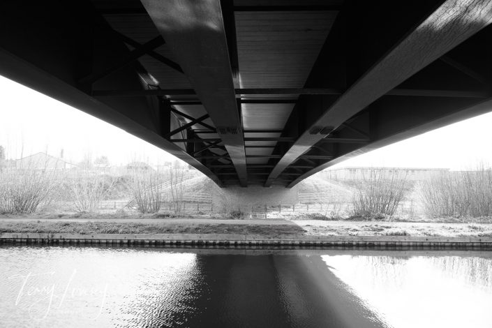 Black and white photo showing the underside of a large metal bridge spanning over a calm river, with light shining through on the grassy bank and leafless bushes in the background.