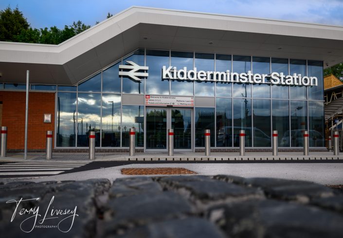 Modern exterior of Kidderminster Station with large glass windows reflecting the sky, bold station name signage, red and silver bollards, and a zebra crossing in front.