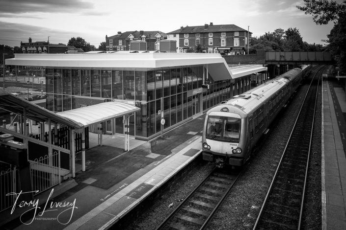 A black and white photo of a modern railway station, with a train stopped at the platform. Glass station building, empty platform, and some houses are visible in the background. Signature Terry Livesey Photography is in the corner.