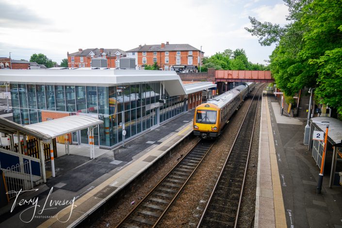 A yellow and grey passenger train approaches a modern station with glass buildings and empty platforms, with brick buildings and trees in the background.
