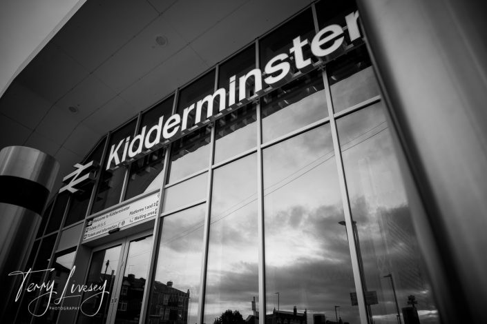 Black and white photo of Kidderminster railway station entrance with large reflective windows showing a cloudy sky. The station sign and railway symbol are visible above the doors.