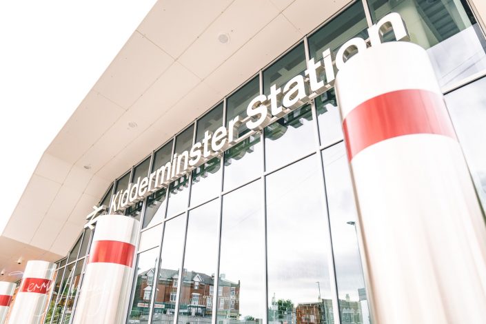 Modern glass exterior of Kidderminster Station with large white sign, reflective windows, and red-striped cylindrical pillars in the foreground. Nearby buildings are visible in the window reflection.