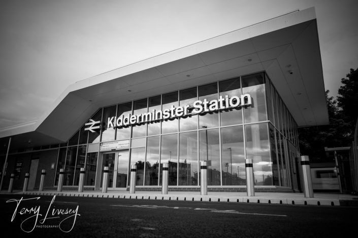 Black and white photo of the modern glass exterior of Kidderminster Station, with large bold signage above the entrance and reflections visible in the windows. The sky is overcast. Photographer’s signature is visible in the corner.