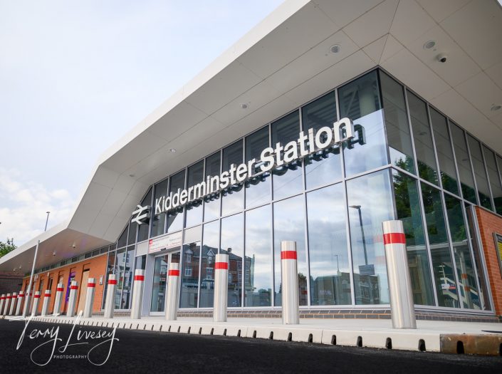 Modern exterior of Kidderminster Station with large glass windows, white roofing, and the station name in bold letters above the entrance. Reflection of nearby buildings visible in the glass.