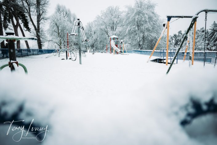A snow-covered playground with swings, climbing frames, and slides, surrounded by trees frosted in snow on a winter’s day. The scene appears quiet and empty.