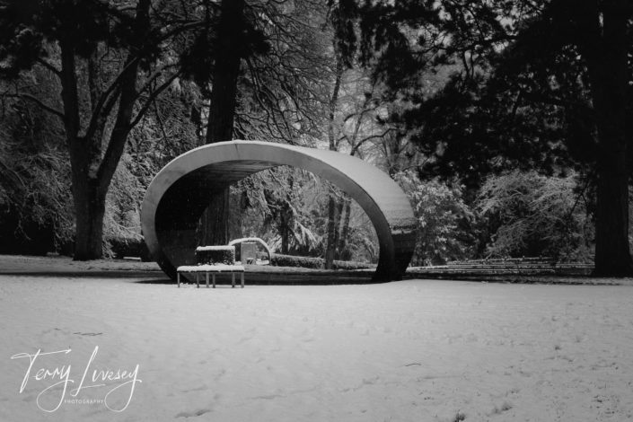 A modern, curved metallic pavilion stands in a snow-covered park surrounded by tall trees. A bench sits nearby, and everything is dusted with freshly fallen snow. The photo is black and white.
