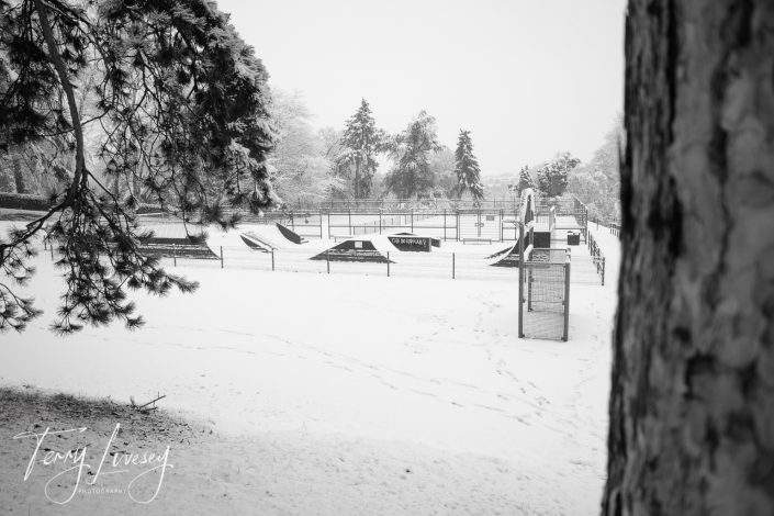 A black and white photo of a snow-covered skatepark surrounded by trees. The park is empty, with ramps and fences visible. Snow blankets the ground, and tree branches frame the scene.