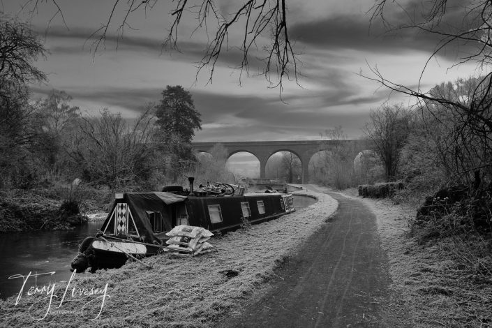 A black-and-white photo of a narrowboat moored along a frosty canal towpath, with leafless trees and an arched stone bridge in the background under a cloudy sky.
