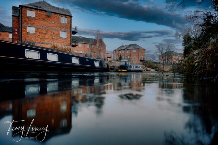 A narrow canal with boats is lined by brick flats at sunset. The water reflects the sky and buildings. Bare trees and clouds are visible, and the image is signed Terry Livesey Photography in the lower left corner.