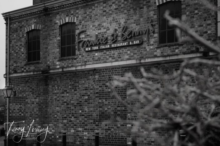 Black-and-white photo of a brick building with a large sign reading “Frankie & Benny’s New York Italian Restaurant & Bar.” Some branches are visible in the foreground, partially obscuring the lower right-hand corner.