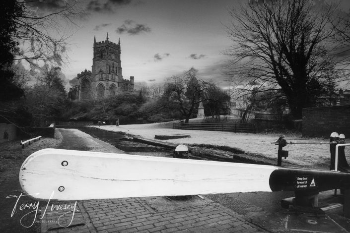 Black and white photo of a canal lock gate in the foreground, with a large historic church and leafless trees in the background. The scene has a tranquil, wintry atmosphere. The image is signed Tony Ivesey Photography.