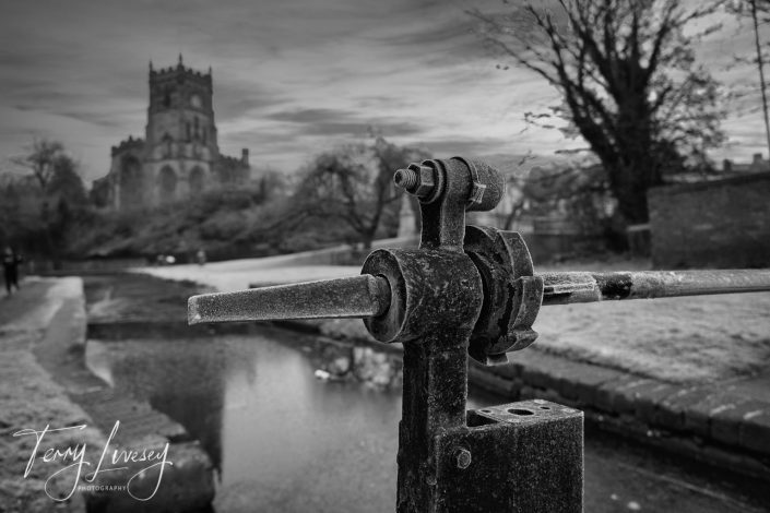 A black-and-white photo shows a close-up of a metal lock gate mechanism by a canal, with a historic church and leafless trees blurred in the background. The sky is overcast, creating a moody atmosphere.