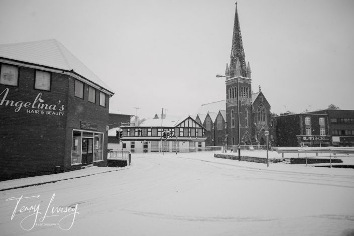 A snowy street scene featuring a church with a tall steeple, a hair and beauty salon called Angelinas, and surrounding buildings, all covered in snow. Tyre tracks are visible on the road.