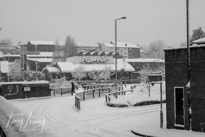 A snowy urban street with buildings and trees covered in snow. A Pizza Hut restaurant is visible in the centre background. Few people are walking, and tyre tracks mark the snow-covered road. Photo credit: Tony Livesey.