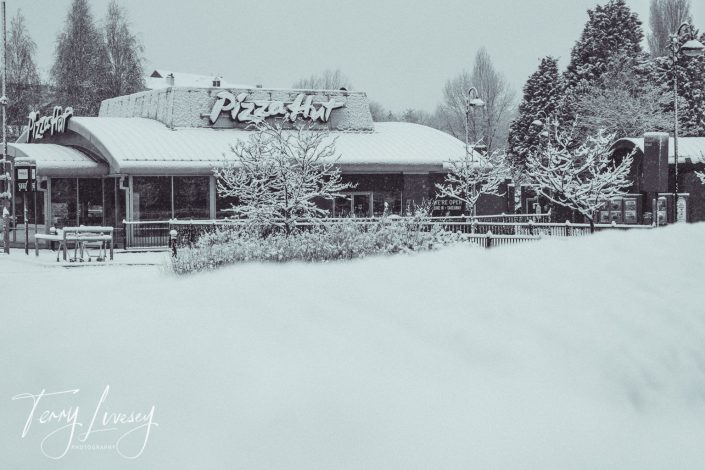 A Pizza Hut restaurant surrounded by snow-covered trees and ground, with snow blanketing the roof and outdoor areas; the scene appears quiet and wintry.