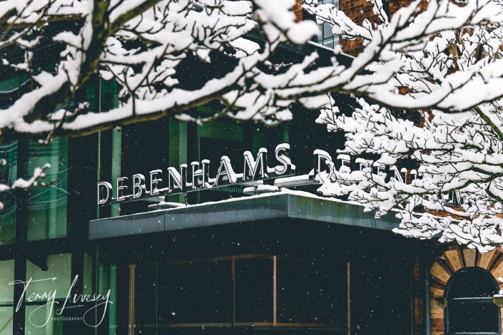 Snow-covered branches partially obscure the Debenhams store sign on a building; snow is gently falling, and the scene has a wintry atmosphere.