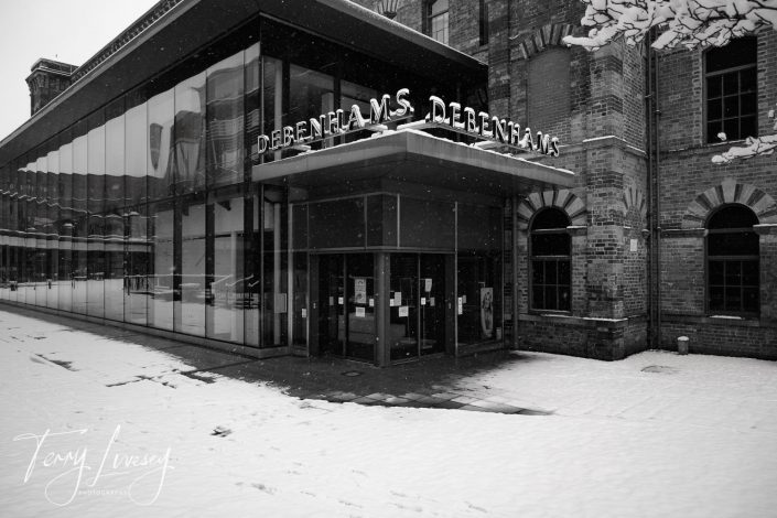 Black and white photo of a Debenhams store with glass walls, attached to a brick building. Snow covers the ground and part of the pavement. Several notices are posted on the entrance doors.