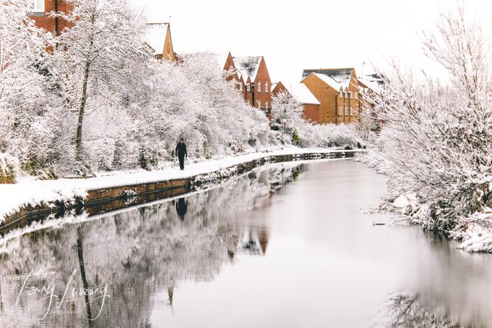 A person walks along a snow-covered path beside a calm canal, with red-brick houses and trees blanketed in snow reflecting in the water on a winter's day.
