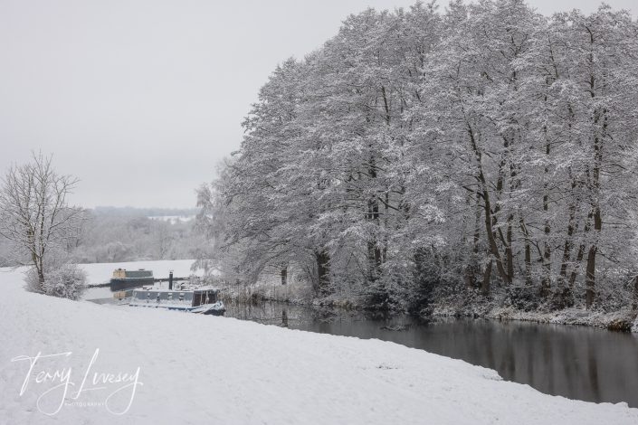 A snowy riverside scene with trees covered in snow, a calm river, and two canal boats moored along the bank. The landscape is peaceful and wintry under an overcast sky.
