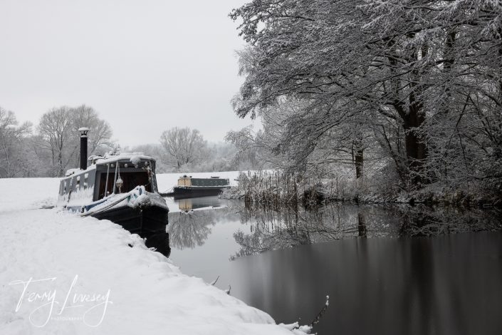 A narrowboat and a barge moored on a calm, snow-covered riverbank, surrounded by bare trees and fields blanketed in snow on a cloudy winter’s day. The water reflects the wintry landscape.