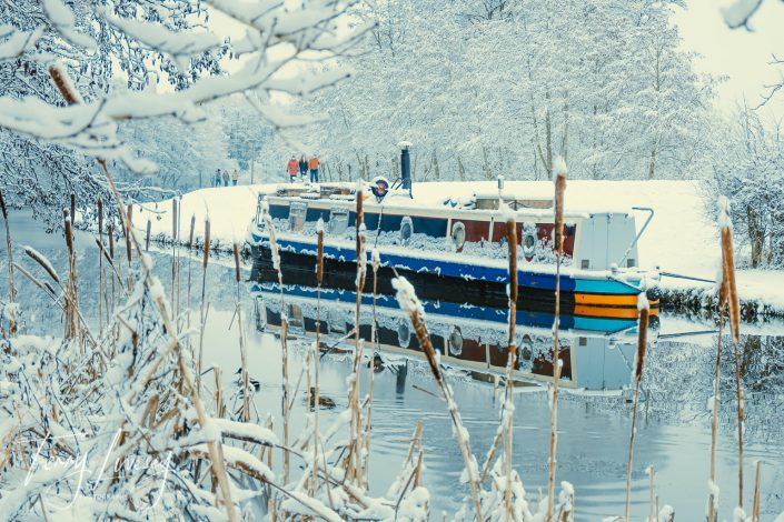 A narrowboat is moored on a snow-covered canal, surrounded by snow-laden trees and reeds. The boat and winter scenery are reflected in the calm water. A few people walk along the snowy path in the background.