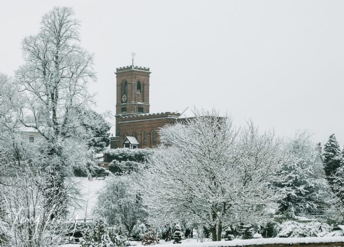 A red-brick church with a clock tower stands surrounded by snow-covered trees and bushes on a wintry day. The scene is peaceful and blanketed in white.