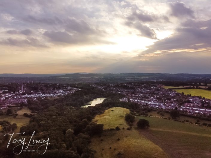 Aerial view of a rural town at sunset, with fields, trees, and a small lake surrounded by houses under a partly cloudy sky. The sun is partially hidden, casting a soft light across the landscape.