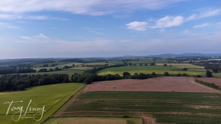 Aerial view of green and brown farmland under a partly cloudy blue sky, with rolling hills and scattered trees in the distance. The image is signed Terry Livesey Photography in the lower left corner.