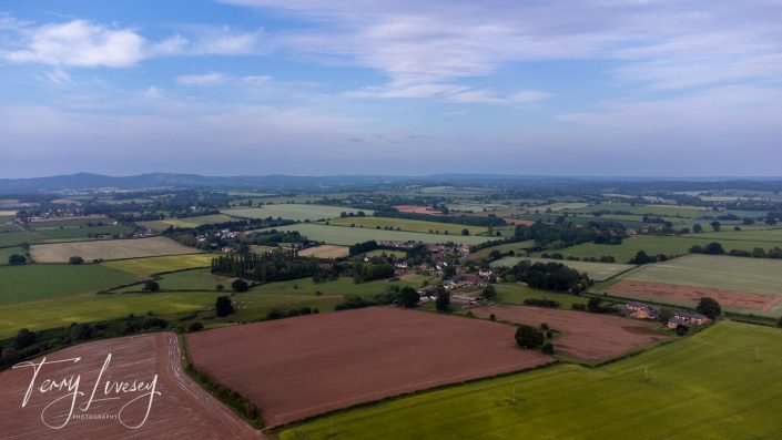 Aerial view of a rural landscape with patchwork fields, small clusters of houses, and distant wooded hills under a blue sky with scattered clouds. A signature reading Terry Livesey Photography is visible in the corner.