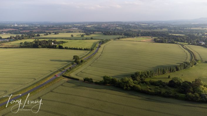 Aerial view of green fields divided by a winding road with a red vehicle. Trees and hedges line the fields, and distant buildings are visible under a partly cloudy sky.
