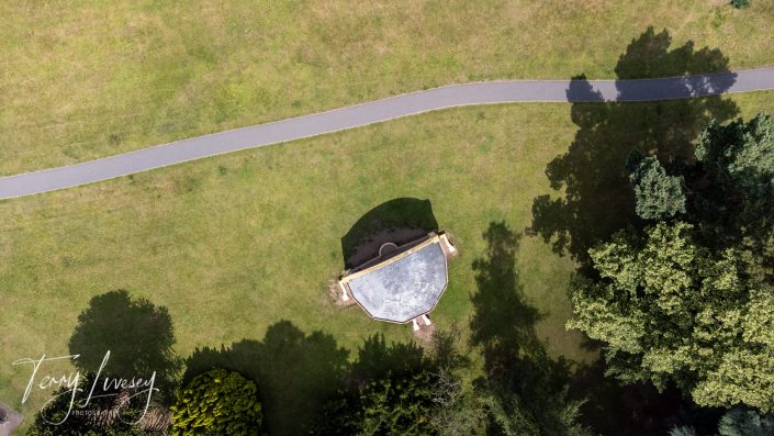 Aerial view of a small hexagonal building surrounded by grass, trees, and a curving paved path in a park setting on a sunny day.