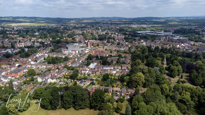 Aerial view of a town surrounded by green trees and fields under a partly cloudy sky. Residential houses, streets, and larger buildings are visible, with open countryside in the background.
