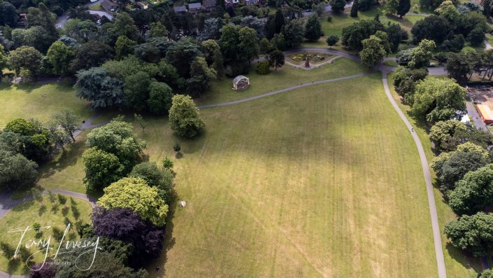 Aerial view of a large, open grassy park surrounded by trees, with footpaths curving through the area, a small bandstand, and a few people visible in the distance. Houses border the park at the top.