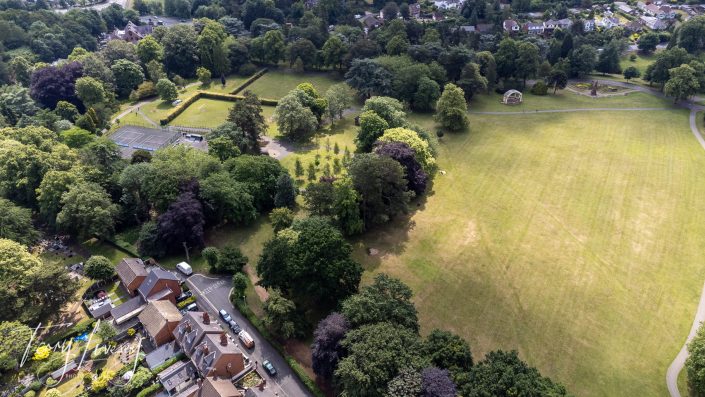 Aerial view of a park with large grassy fields, scattered tall trees, a tennis court, a bandstand, and nearby residential houses along a tree-lined road.
