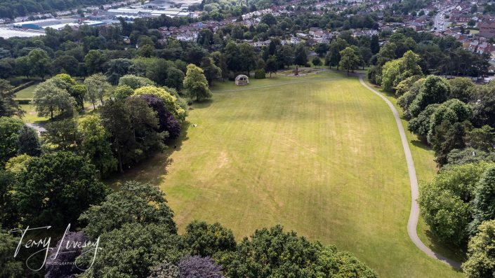 Aerial view of a large grassy field bordered by trees, with a path curving around the right side. A few structures are visible on the grass, and a residential area is in the background.