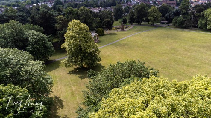Aerial view of a large green park with scattered trees, walking paths, a small performance pavilion, and some benches. Houses and more trees are visible in the background.