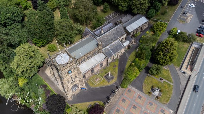 Aerial view of a historic stone church surrounded by trees, car parks, and roads. The church has a tower, pitched roofs, and a courtyard with pathways. Cars are parked nearby. The photo is signed by Terry Livesey.