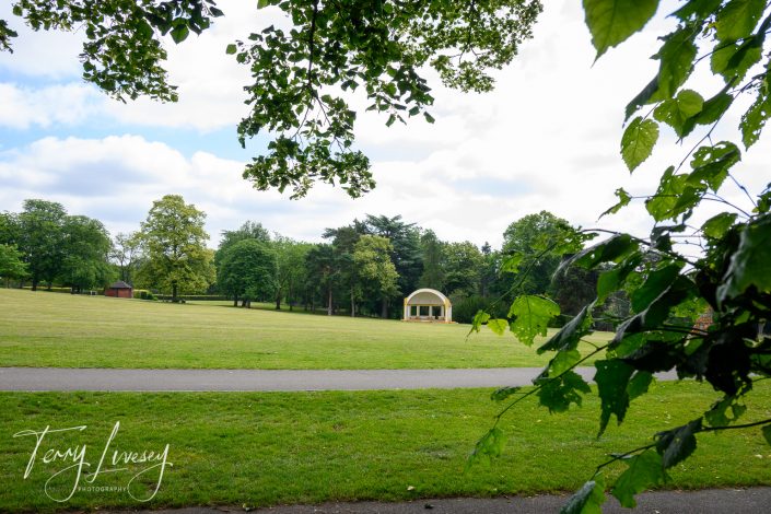 A green park with scattered trees, a paved path, and a small domed bandstand in the distance, viewed through leafy branches on a bright, partly cloudy day.