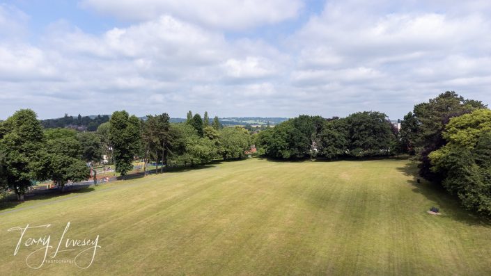 A wide, grassy field bordered by trees under a partly cloudy sky, with a park and distant hills visible in the background. The image features the signature “Terry Livesey Photography” in the lower left corner.